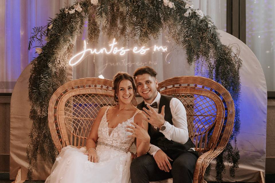 Pareja sentada frente a un arco de plantas enseñando sus anillos